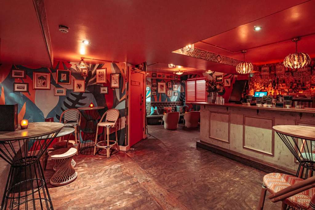 Main room with red ambient lighting, bar counter, high-top tables, decorative artwork, and pendant lighting fixtures.