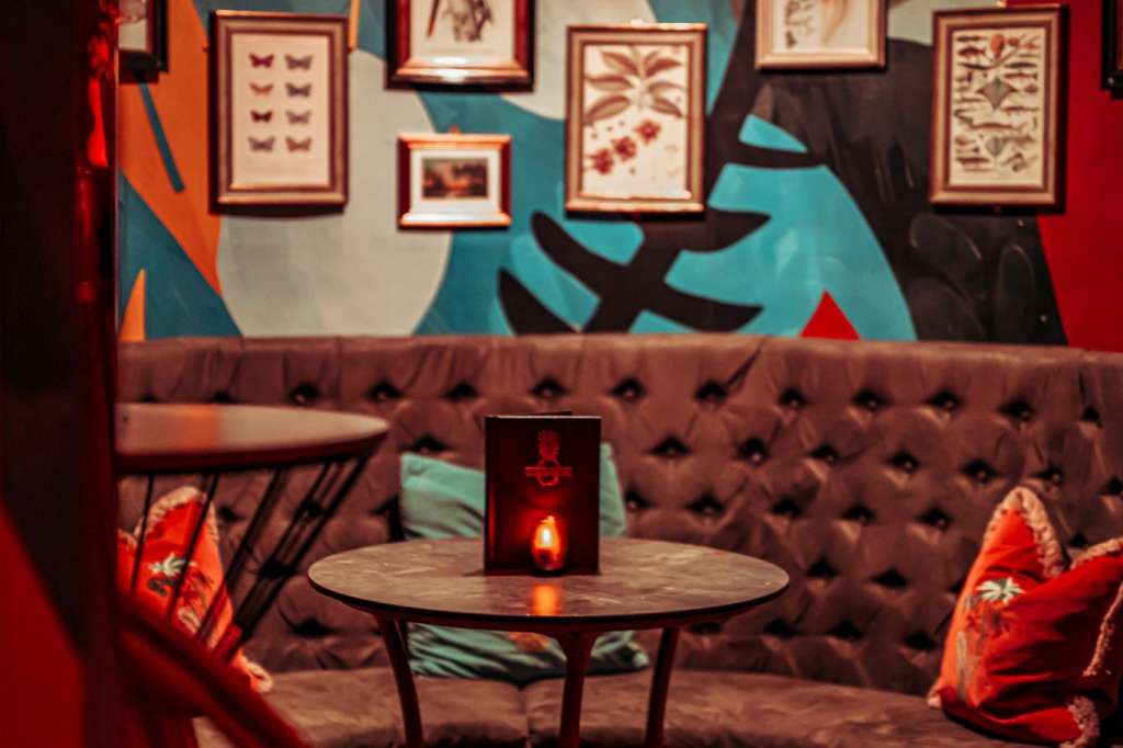 Main room with red ambient lighting, tufted seating, round table, and framed botanical artwork on walls.