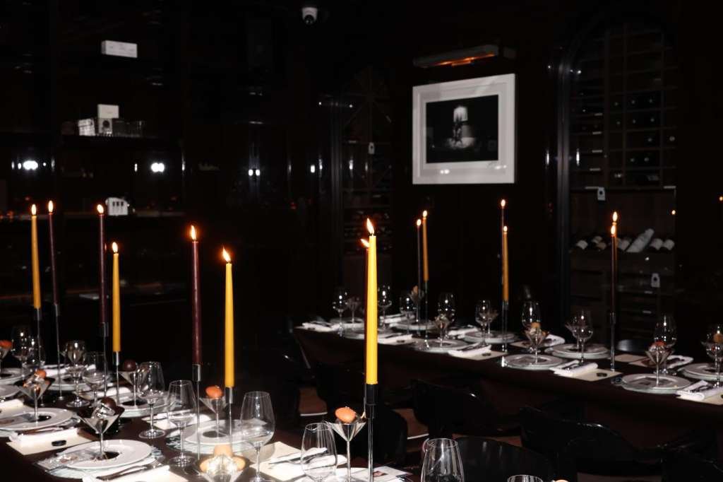 Elegant dining table set with white plates, gold candlesticks, glassware, and dark linens in a modern black interior.