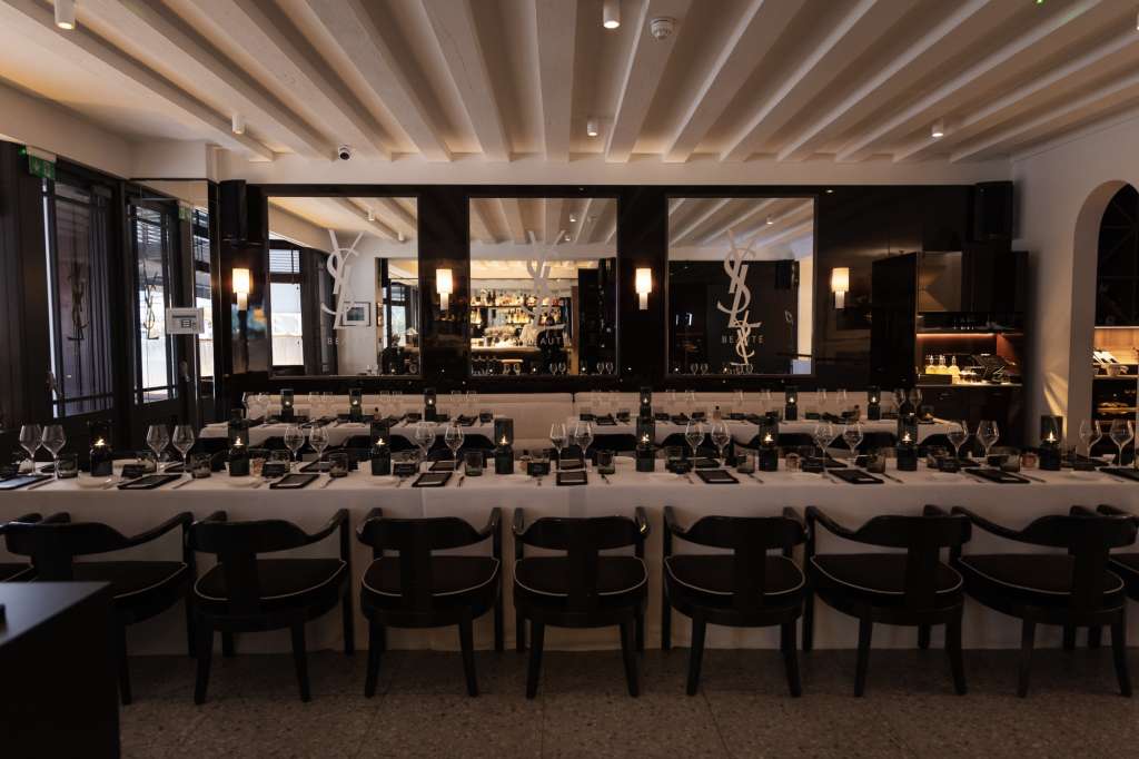 Long dining table set with place settings, glassware, and black chairs facing a mirrored bar with ambient lighting.
