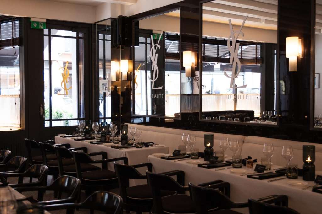 Modern dining room with long tables, black chairs, white linens, candlelight, and floor-to-ceiling windows.