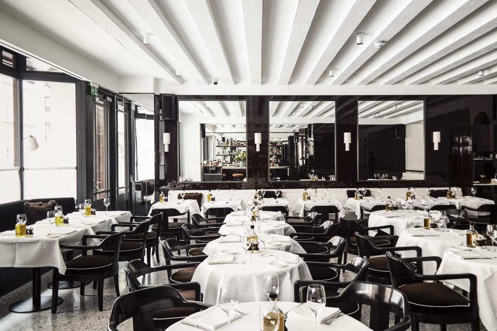 Fine dining restaurant with round tables, white linens, black chairs, and modern ceiling beams.