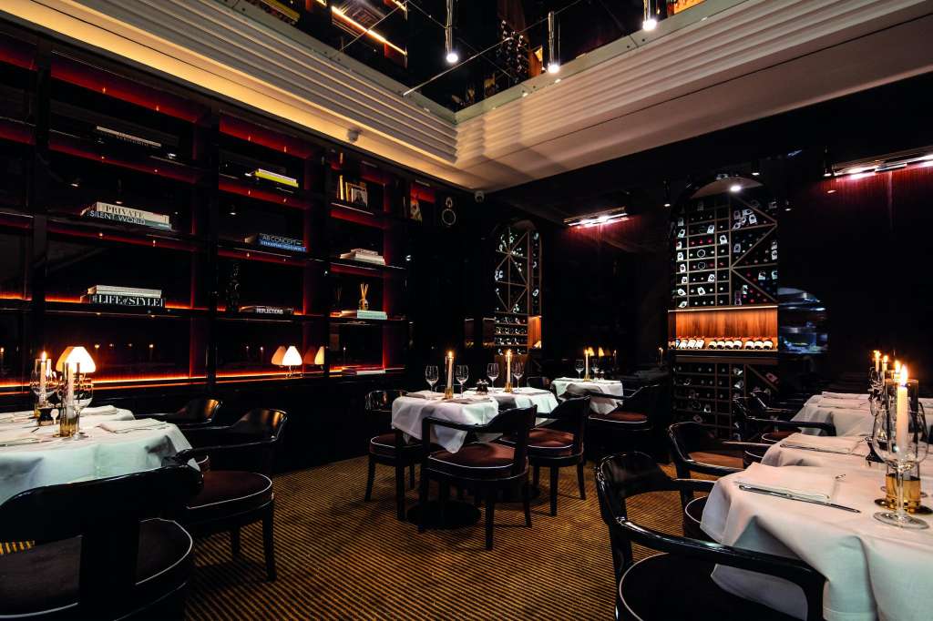 Fine dining room with round tables set with white linens, black chairs, and ambient lighting from illuminated wall shelving.