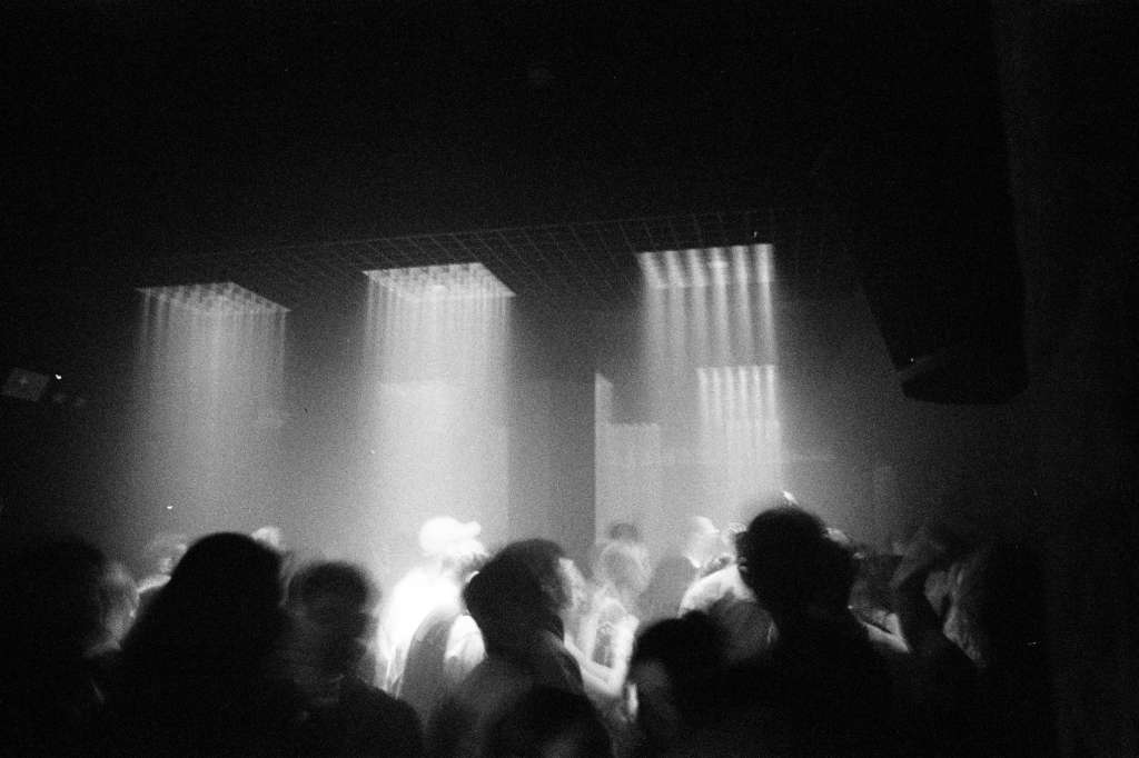 Crowd silhouetted against dramatic vertical light beams in a darkened venue space.