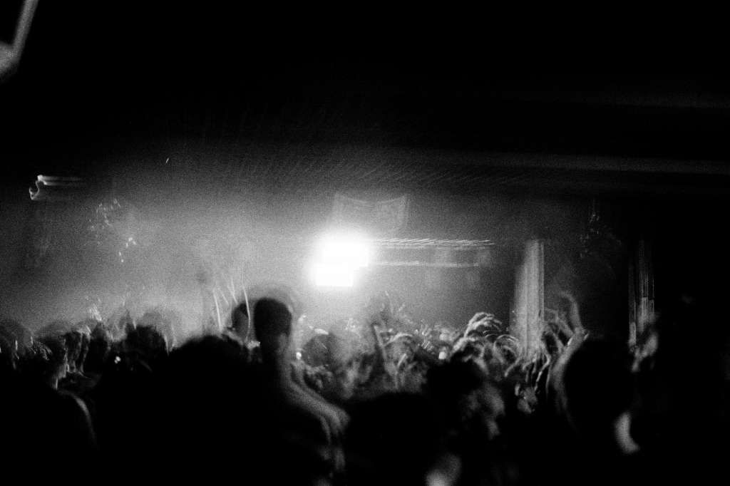 Crowded indoor venue with stage lighting and raised hands in black and white.