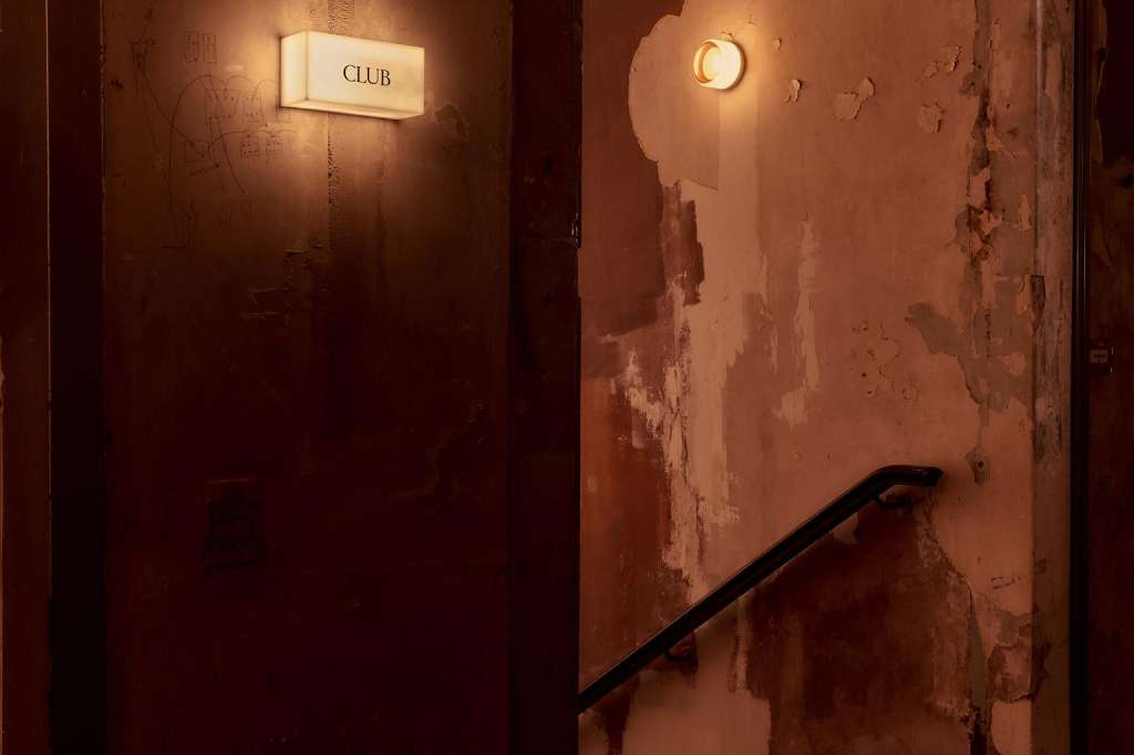 Dimly lit corridor with weathered walls, illuminated signage reading 'CLUB', and a black handrail.