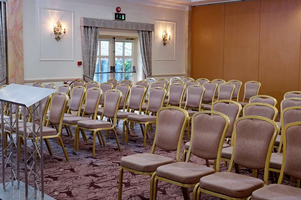 Meeting room with rows of gold-framed chairs facing a small podium, elegant wall sconces, and glass doors overlooking grounds.