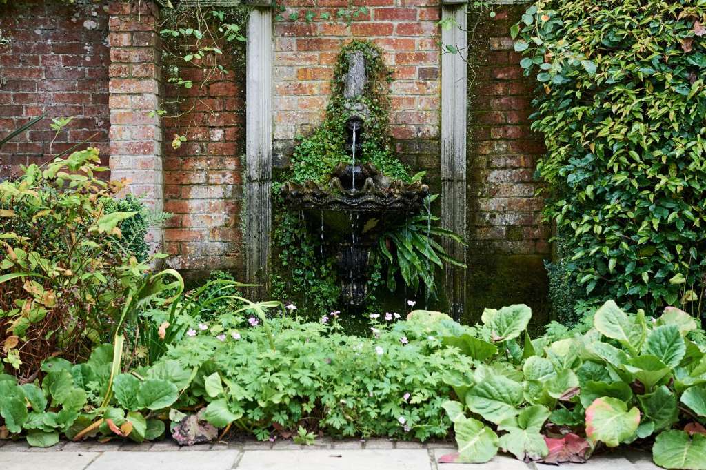 Brick courtyard with ivy-covered walls, ornamental fountain, and lush green plants in foreground.