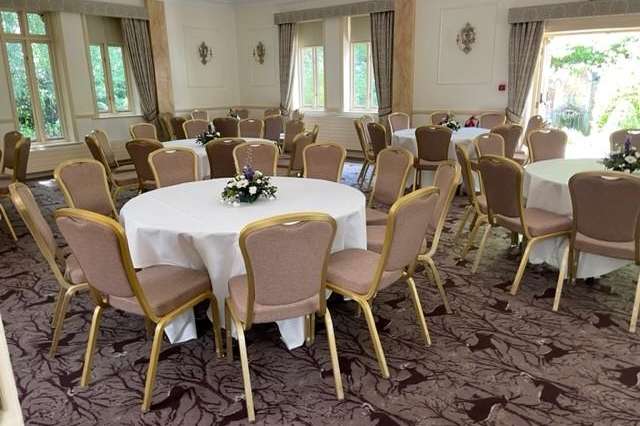 Ballroom set with round tables, white linens, upholstered chairs, and floral centerpieces. Large windows and elegant drapes.