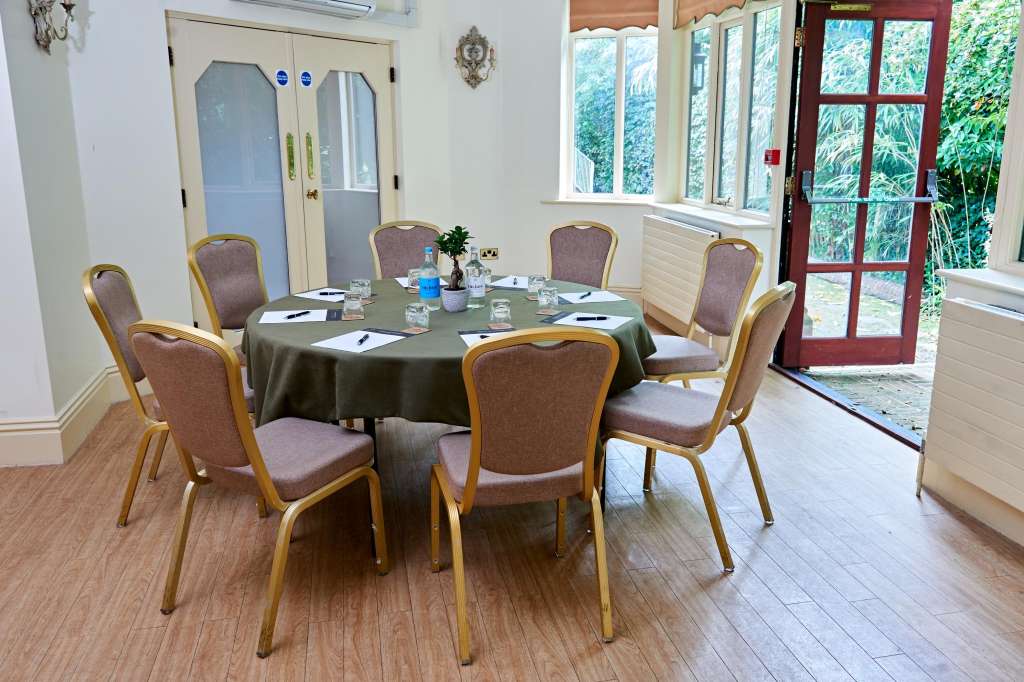 Meeting room with round table dressed in olive cloth, six upholstered chairs, water glasses, and notepads. Large windows overlook gardens.