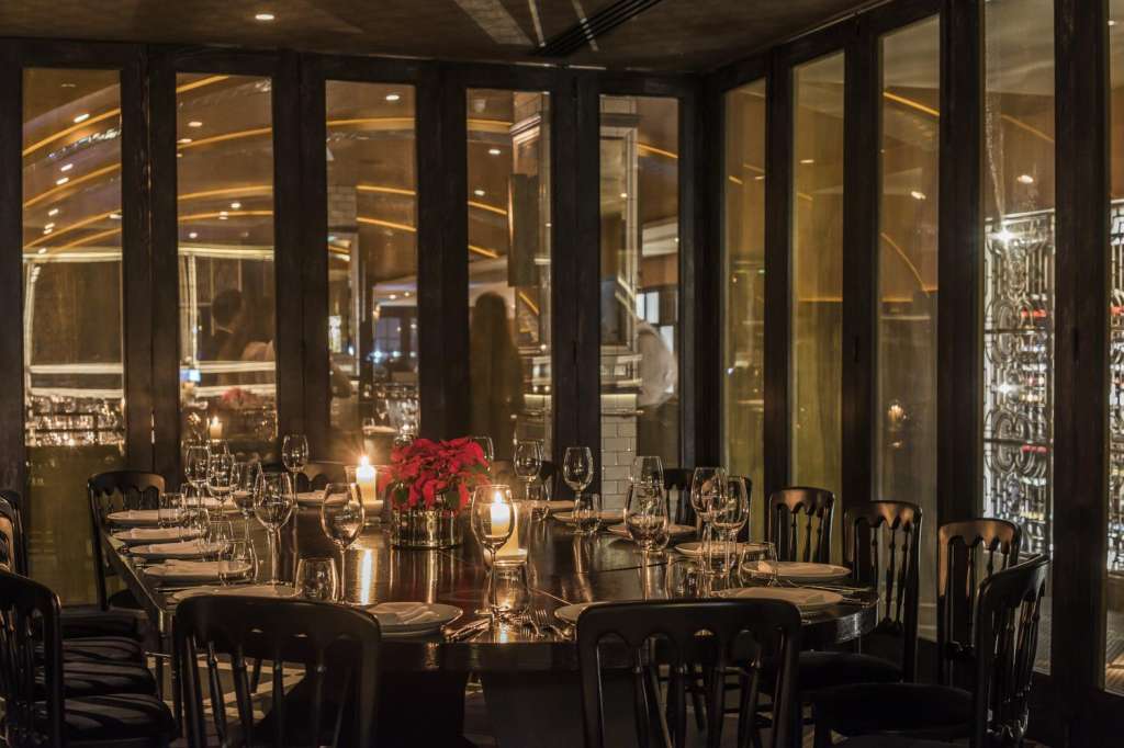 Dining room set for a formal seated dinner with long table, place settings, wine glasses, red floral centerpiece, and floor-to-ceiling windows.