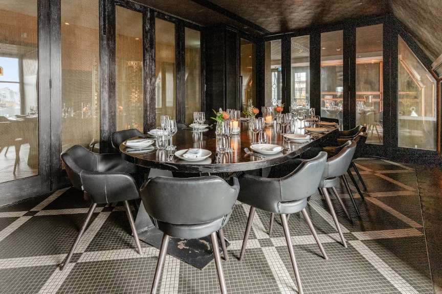 Modern dining room with curved black table, grey chairs, place settings, and floor-to-ceiling windows overlooking the city.
