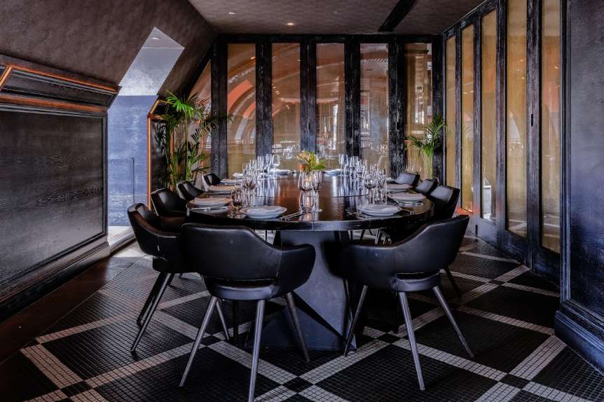 Modern dining room with round table, black chairs, place settings, and floor-to-ceiling windows overlooking landscape.