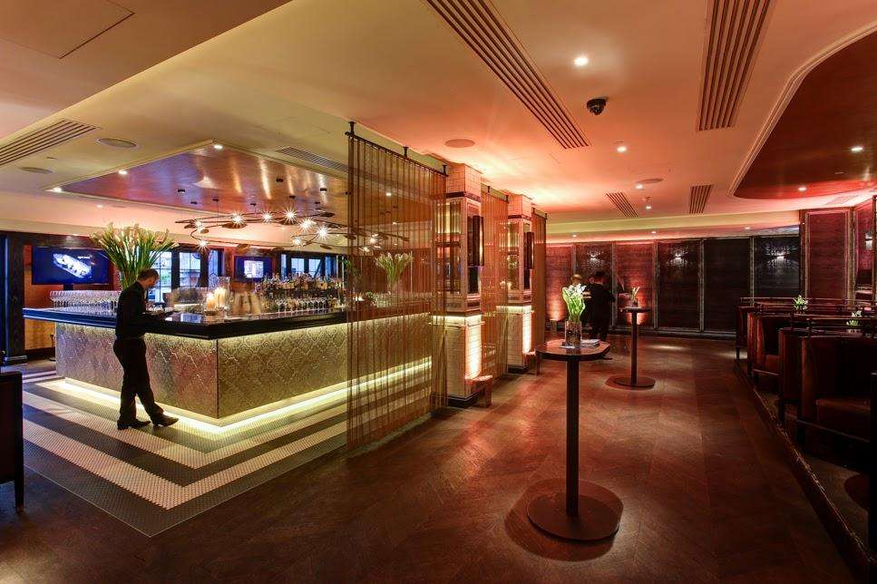 Modern bar and lounge with illuminated counter, ambient lighting, high ceilings, and standing figure near bar.
