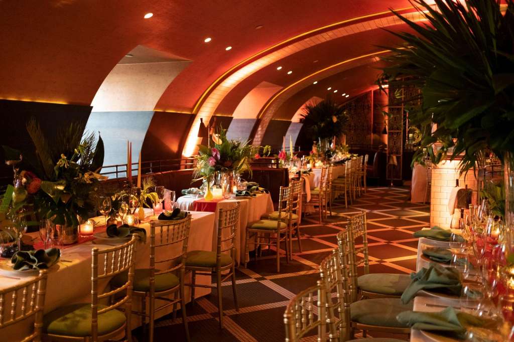 Curved dining room with arched ceiling, round tables set with linens, floral centerpieces, and ambient lighting overlooking water.