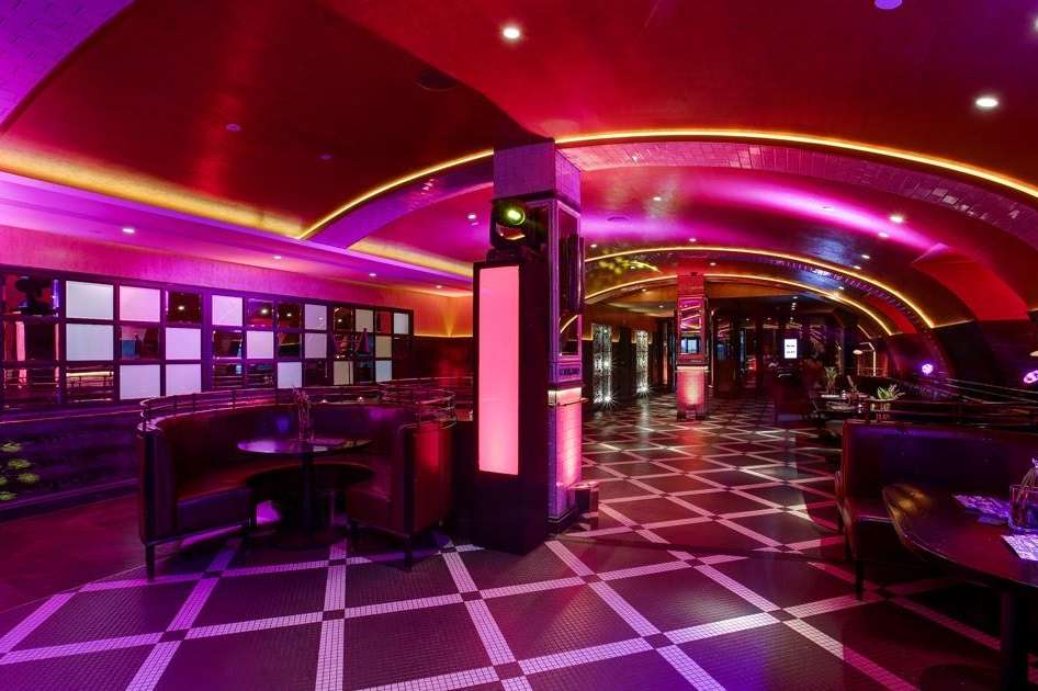 Bar and lounge space with vibrant pink and purple ambient lighting, curved ceiling, geometric floor, and modern furnishings.