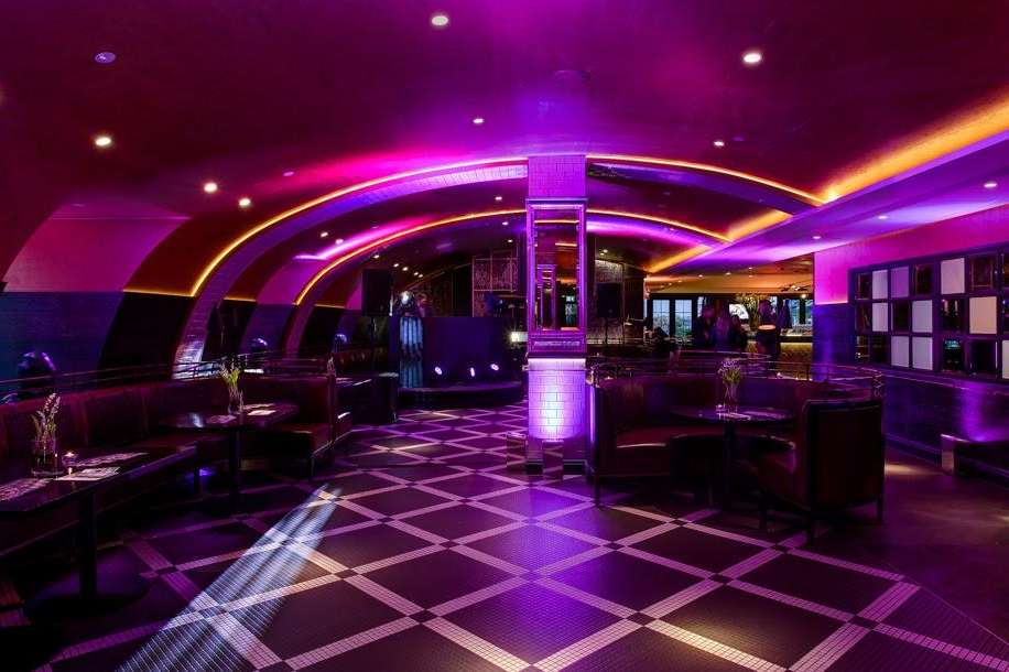 Modern bar and lounge with purple and gold ambient lighting, curved architectural details, geometric floor pattern, and contemporary furnishings.