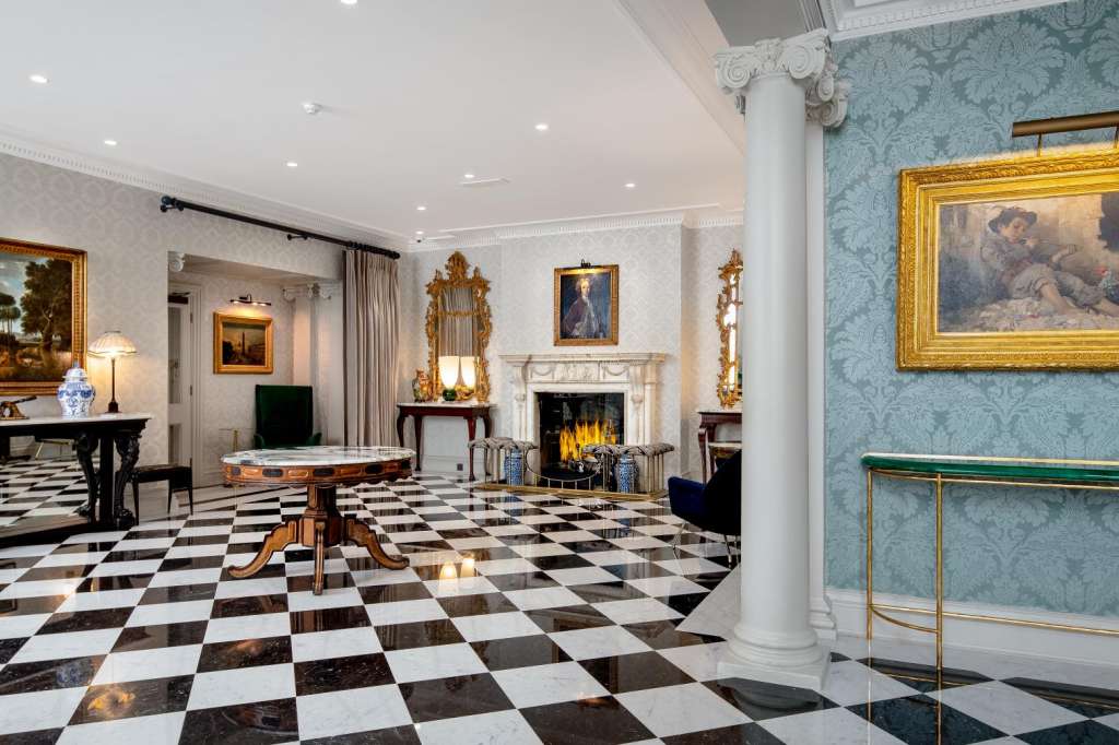 Grand reception room with black and white checkered marble floor, fireplace, ornate gold-framed paintings, and classical columns.
