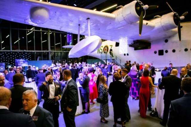Large aircraft hangar with guests in formal attire mingling under purple ambient lighting.