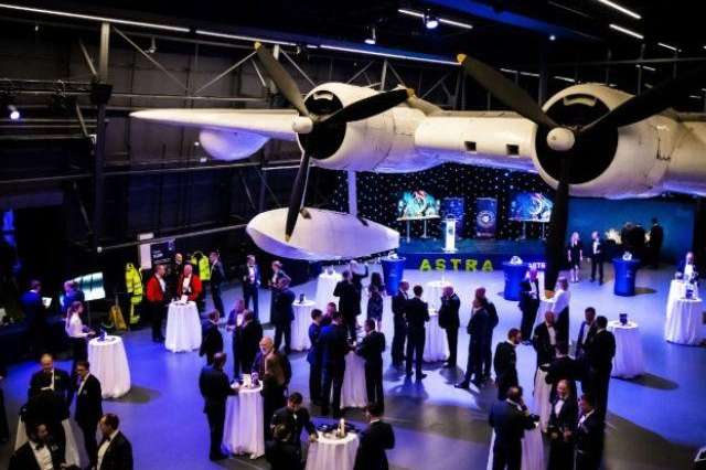 Large hangar with vintage aircraft overhead, blue lighting, guests in formal attire mingling around high-top tables.