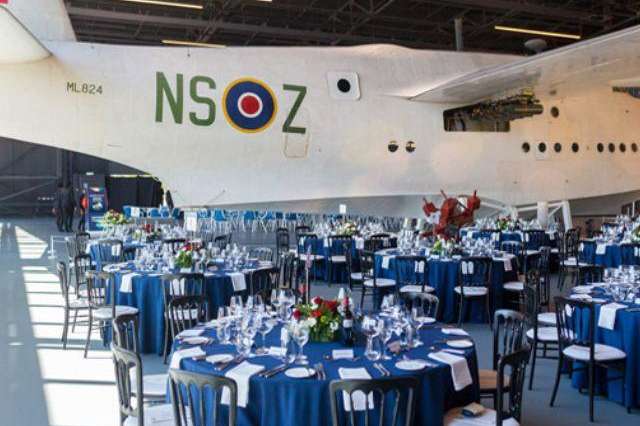 Aircraft hangar interior with round tables dressed in blue linens, place settings, and floral centerpieces.