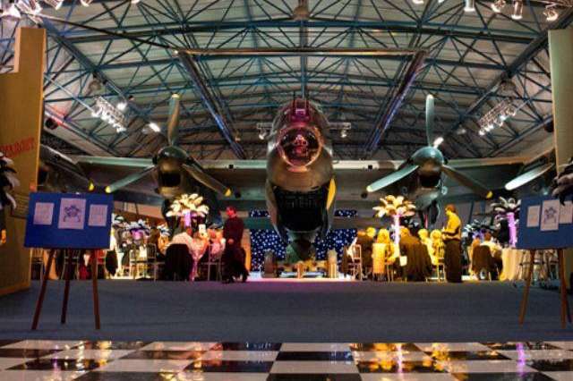 Large hangar with vintage aircraft suspended above, illuminated stage with decorative lighting and displays below.