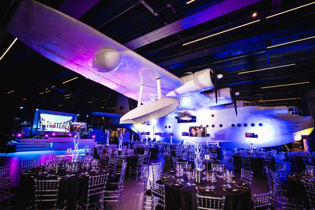 Sunderland Hall set up for a formal dinner with round tables, dramatic purple and blue lighting, and aircraft display overhead.