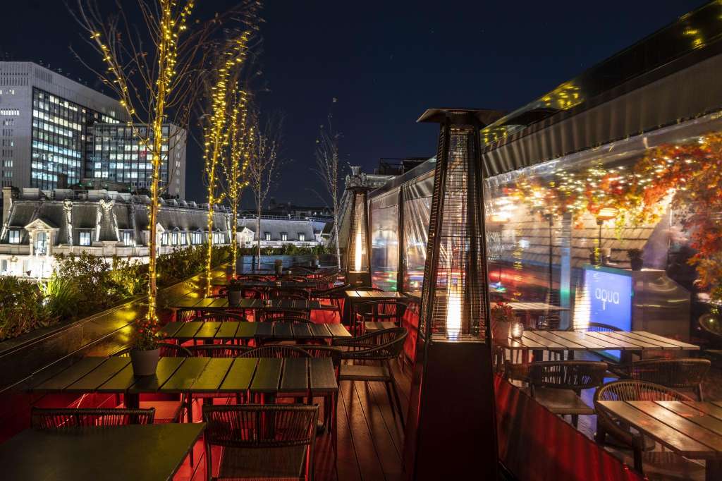 Rooftop terrace at night with red seating, illuminated trees, and city skyline views.