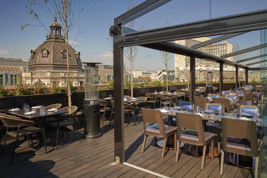 Rooftop terrace with multiple dining tables, woven chairs, and city skyline views. Bare trees and architectural dome visible.