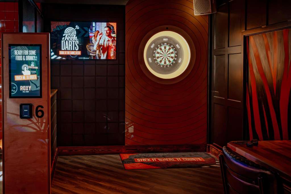 Games room with illuminated dartboard, red wood paneling, digital screens, and branded signage.