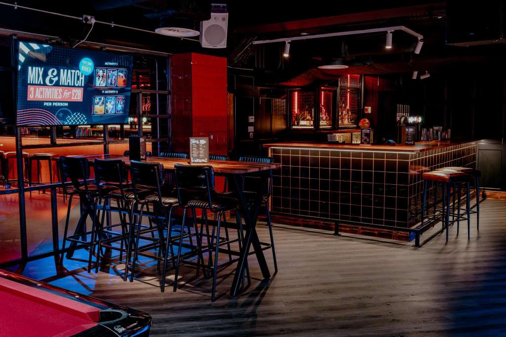 Modern bar and lounge space with red and blue neon lighting, high-top tables, bar counter, and gaming screens.