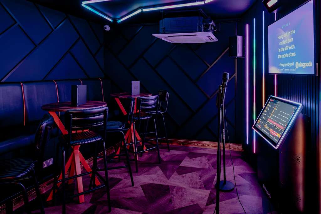 VIP Room with neon-lit bar seating, high-top tables, geometric wall paneling, and projection screen displaying event information.