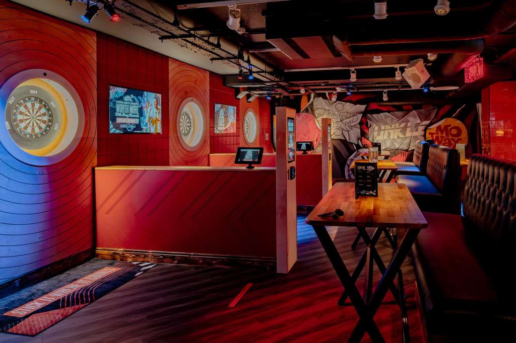 Karaoke room with red walls, illuminated dartboards, high-top tables, and neon signage under colored lighting.