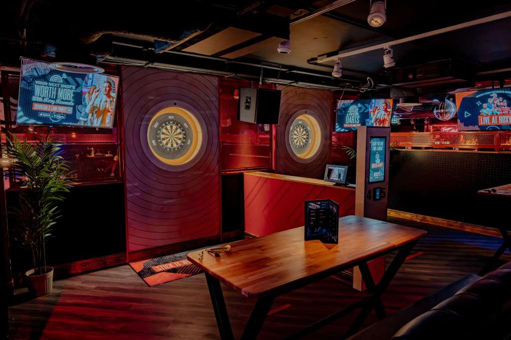 Karaoke room with two illuminated dartboards, wooden table, red and purple lighting, and neon signage.