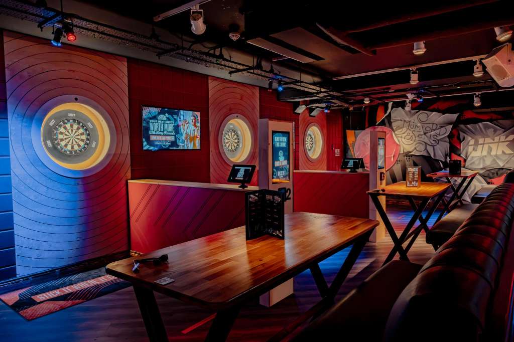 Karaoke room with dartboards, high tables, red walls, blue and red lighting, and casual seating.