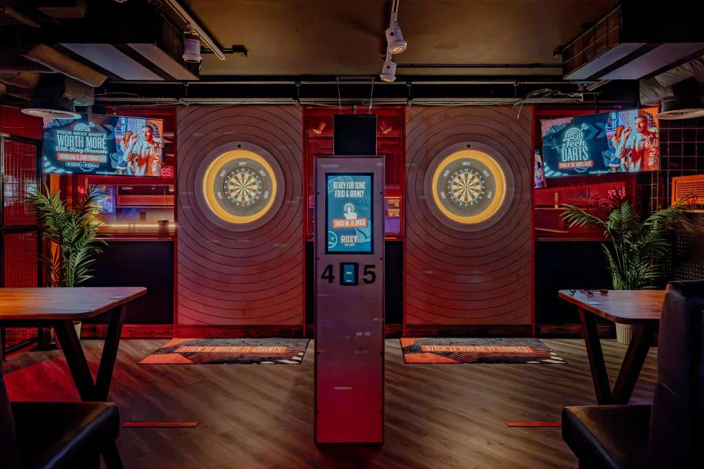 Karaoke room with two illuminated dartboards, red walls, tables, and branded signage.