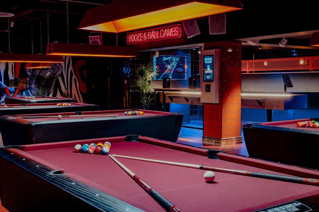 Pool tables with colorful balls and cues in a neon-lit games room with booze and ball games signage.