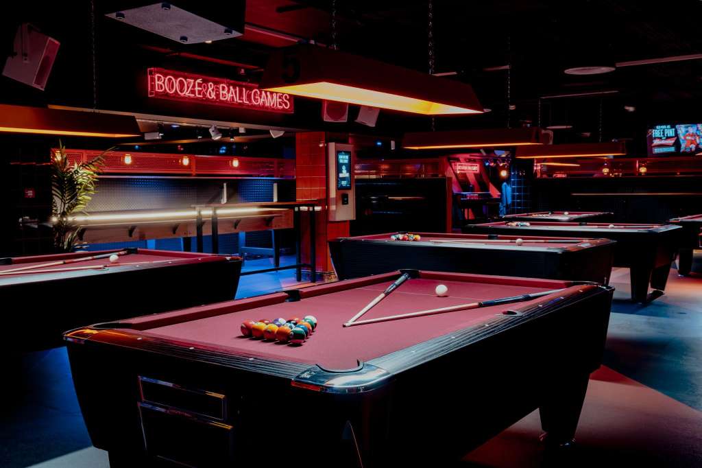 Pool tables with red felt and neon signage in a dimly lit games room with blue flooring.