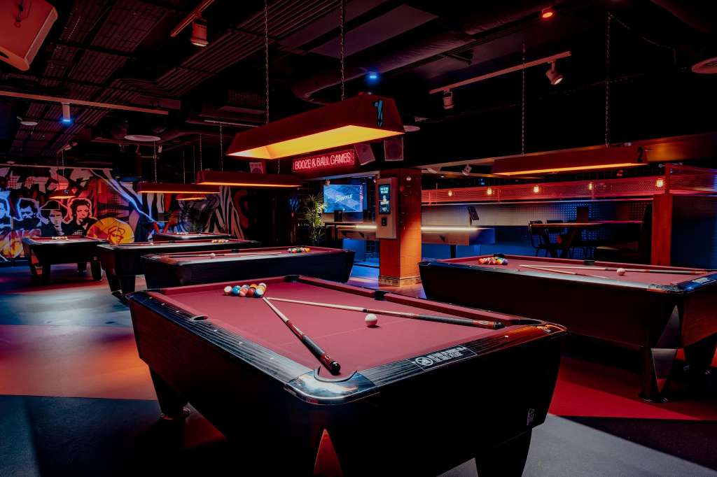 Games room with multiple pool tables, neon lighting, and arcade machines in red and blue tones.
