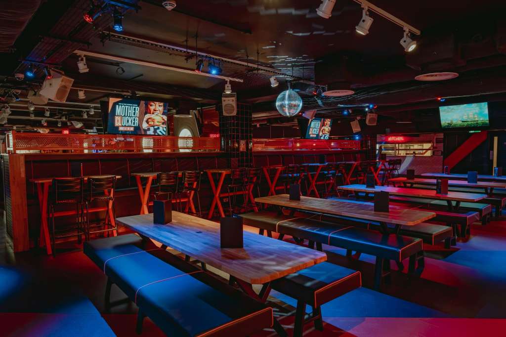 Gaming hall with bowling lanes, red bar seating, blue and red lighting, disco ball, and video screens.