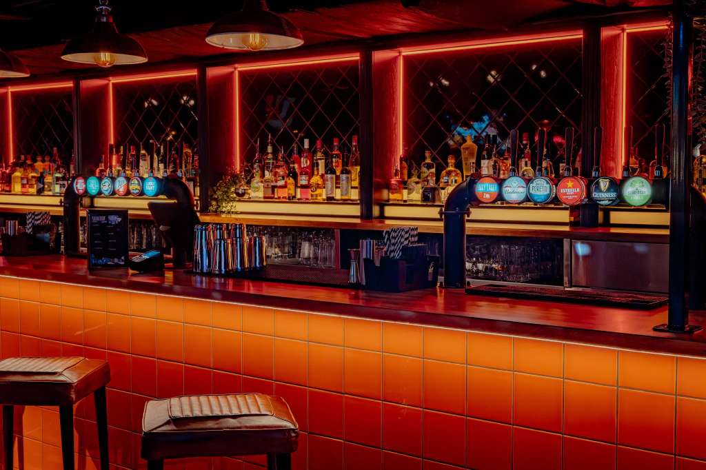 Bar with red neon lighting, mirrored shelving, spirit bottles, and colorful tap handles. Staff member visible behind counter.