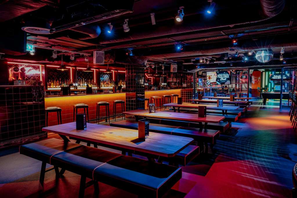 Gaming hall with ping pong tables, bar seating, neon lighting in red and blue, and arcade games.