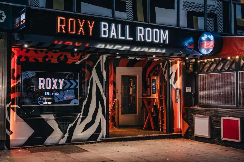 Exterior storefront of Roxy Ball Room with neon signage, red and black branding, and glass entrance doors at night.
