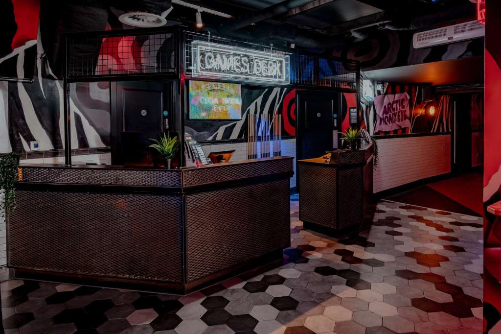 Karaoke room with neon signage, gaming booths, hexagonal flooring, and red ambient lighting.