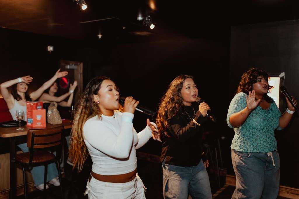 Group of people singing karaoke in a dark venue with stage lighting and bar seating visible in background.