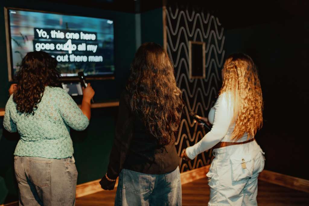 Three people playing karaoke in a dark room with a screen displaying lyrics and geometric wall decor.