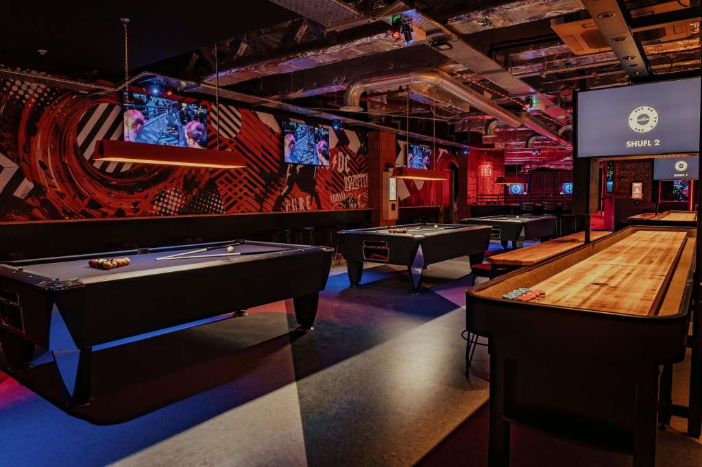 Crazy Pool room with multiple pool tables, shuffleboard, red and black geometric wall graphics, and blue accent lighting.