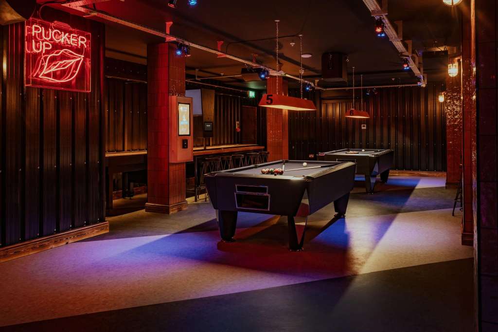 Gaming hall with pool tables, neon signage, red pillars, and colored floor lighting.