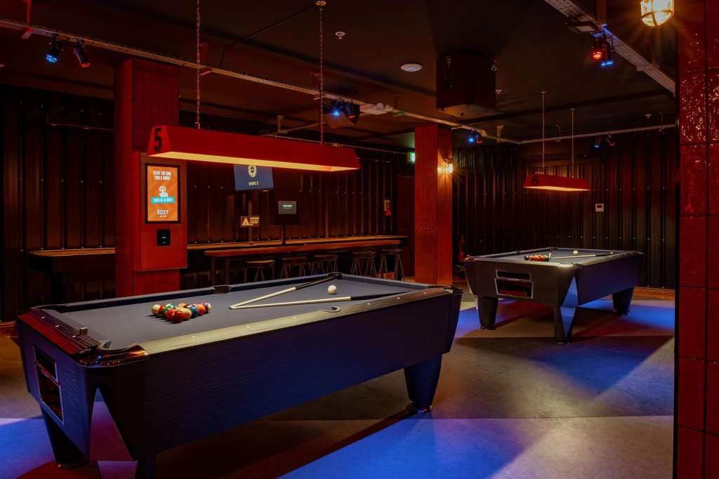 Gaming hall with pool tables, neon lighting, red walls, and bar seating in background.