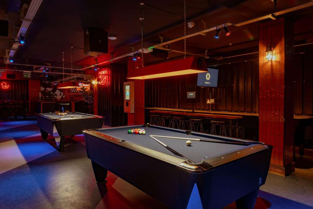 Gaming hall with pool tables, neon signage, red and blue ambient lighting, and bar seating along the back wall.